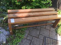 Front and side view of wooden outdoor bench showing three slats and leg detail on stone patio surrounded by plants.