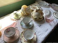 Overview of assorted tea cups, saucers, plates on white cloth, showing floral patterns and gold trims.