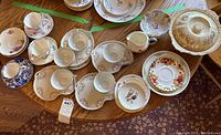 Top-down view of assorted fine china teacups, saucers, plates, creamers, and a covered serving dish arranged on wooden table.