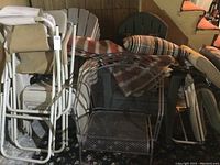 Photo showing folded white metal frame patio chairs with beige fabric seats, a metal mesh chair, green plastic chairs, and several cushions piled on top.
