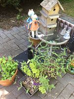 View showing several garden pots with living plants, the small round glass-top table, ceramic cat and frog figurines, wooden multi-compartment birdhouse, and a black folding metal plant stand.