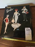 Closed black vintage Barbie carrying case featuring three illustrated Barbie figures in different outfits and a tape measure for scale about 12 inches.