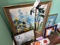 Overview of larger framed landscape painting leaning against wall with other smaller framed pieces stacked beside it.