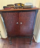 Front view of vintage wooden hall cabinet showing two doors, decorative wood trim and turned legs with small caster wheels. Various small items rest on top.