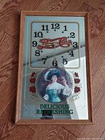 Front view of the oak framed Pepsi-Cola themed wall clock showing clock face, mirrored background, and decorative antique style image.