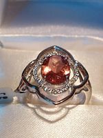 Close-up view of a rhodium plated sterling silver ring with an oval synthetic Zultanite gemstone with a peachy-pink color, surrounded by small clear stones in a floral-inspired setting, displayed on white fabric.