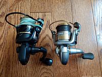 Top view of Fanatik Pirat 2000 and Okuma Avenger AV 306 reels on wooden floor showing spool and handle orientation.