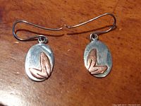 Front view of pair of sterling silver oval earrings with copper leaf accents on a wooden surface