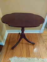 Front view of the vintage occasional table shows the wooden scalloped top and three slender curved legs with castors standing on a hardwood floor.