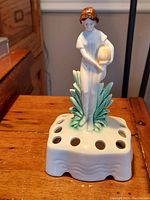 Full front view of the vintage German porcelain flower frog, showing the maiden figure holding a basket above a base with multiple flower holes.