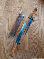 Photo of the vintage meat carving knife, fork, and decorative carved wooden sheath with metal fittings laid on wooden floor.