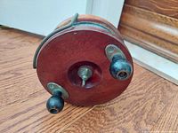 Close-up image of one side of the wooden vintage Peetz salmon fishing reel, showing two black knobs and metal attachments on a wooden surface.