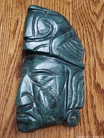 Side profile view showing the carved face with defined nose, lips, and ear ornament of the jadeite Aztec warrior sculpture against a wood background.