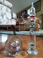 Photo showing vintage Galileo thermometer standing upright with multiple floating colored spheres inside, next to a clear glass spherical paperweight with internal bubbles, both on wooden floor with furniture in background.