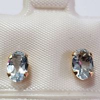 Close-up front view of two oval aquamarine stones set in four-prong 18K gold mountings on white display pad