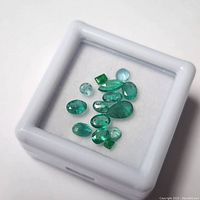 Top-down view of multiple natural emerald gemstones in assorted cuts inside a white display box on white foam