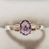 Close-up of a silver ring with an oval faceted amethyst stone, showing the prong setting and polished band.