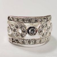 Close-up photo of a wide sterling silver band ring set with clear cubic zirconia stones arranged in multiple rows and circular cutouts, polished finish.
