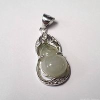 Close-up photo of a silver pendant with a light green carved jade stone and small embedded clear stones on the silver frame.