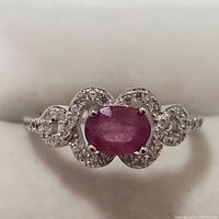 Front view of silver rhodium plated ring featuring an oval 1 ct ruby center stone and surrounding clear stones in a swirl design.