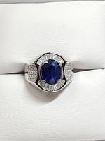 Front view of men's silver ring with central oval blue sapphire gemstone surrounded by baguette cubic zirconia and pavé-style round cubic zirconia on sides.