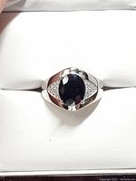 Close-up photo of silver ring with oval dark blue sapphire and cubic zirconia side stones on a white cushion.