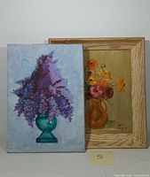 Two oil paintings side by side. Left is a canvas with purple lilacs in teal vase. Right is a framed board painting of colorful flowers in amber pitcher.