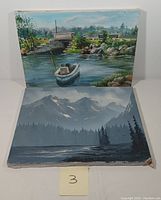 Two oil paintings on canvas displayed side by side. One depicts a boat on a calm waterway with a stone bridge and houses in the background. The other features a misty mountainous scene with pine trees and a lake foreground. Both have visible texture from the paint and some wear on canvas edges.