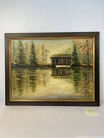 Framed oil painting showing a lakeside home with trees reflected in water, signed by artist, approximately 21 3/4 by 27 1/2 inches.