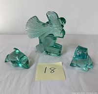 Three green art glass sculptures featuring a bulldog, dolphin and Aztec figure together on a white surface with a yellow sticky note labeled '18'.