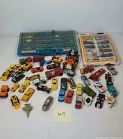 Overview photo of approximately 45 miniature die-cast toy vehicles with various types such as cars, trucks, construction equipment, military vehicles, and a small fighter jet, along with a Mickey Mouse figure in a vintage car and two storage cases.