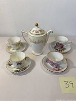 Front view of the vintage porcelain Minton teapot surrounded by four collectible porcelain teacups and saucers with various floral designs and gold accents, all arranged on a white surface.