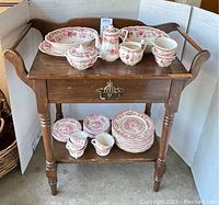 Photo of antique wood wash stand with drawer and towel rods, showing red floral pattern transferware dishes on top and lower shelf with plates and cups.