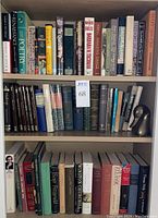 Three shelves filled with books of various genres and conditions, including classics, poetry, and biographies, with a brass duck head bookend on the middle shelf.