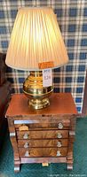 Brass lamp with cream pleated shade sitting on top of the solid wood side table in front of a plaid wallpaper.