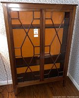 Front view of English antique wooden bookcase with glass doors showing geometric inlay and seller's auction tags.
