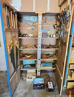 Wide shot of tall wooden cabinet open showing shelves with saws, hammers, screwdrivers, clamps and other tools, plus a large spirit level standing vertically.