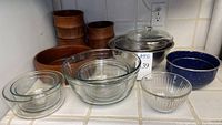 Set of kitchen bowls arranged on countertop including glass, wooden, enamel, and aluminum pieces with glass lid