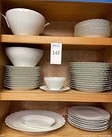 Shelves displaying the porcelain dinnerware set including plates, bowls, tureen and serving plates, all white with raised texture and scalloped edges.