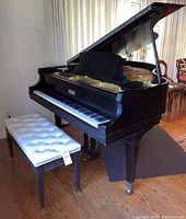 Front right three-quarter view of black grand piano with lid open and white leather bench in front