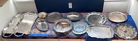 Wide view of all 13 silver and silver tone serving trays and platters arranged on a surface showing variety of shapes and sizes.