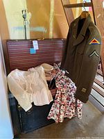 Black steamer trunk open showing interior tray and clothes including white blouse, floral skirt, hat, and ballet flats, with wool army jacket hanging nearby.