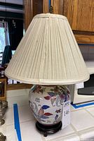 Front side view of ceramic vase-shaped table lamp with cream pleated shade, showing colorful leaf pattern and wooden stand