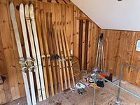 Six pairs of vintage wooden cross country skis leaning against wall with four pairs of ski poles and a pair of vintage snowshoes visible on floor, various accessories scattered on floor.