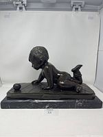 Side view of bronzed statue showing boy playing with ball on marble base, highlighting the detailed textured surface and posture.