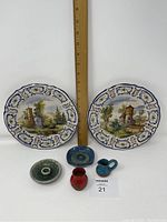 Front view of two French faience hanging plates with scenic village designs, two miniature vases (red and blue), and two ceramic candle holders with green glaze. A ruler is placed for scale.