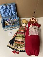 Photo showing the vintage woven sewing basket lined with blue satin and filled with sewing supplies alongside two ethnic costumed fabric dolls lying flat.