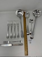 Overview of silver plated serving pieces with ruler for scale, showing various ladles, forks, and cutting utensils in fiddle and old english patterns.