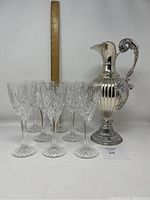 Front view of silver plated wine ewer next to eight pressed crystal wine glasses with ruler showing height