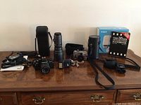Wide shot showing entire lot including cameras, lenses, flash, binoculars, carrying cases, and book on a wooden table